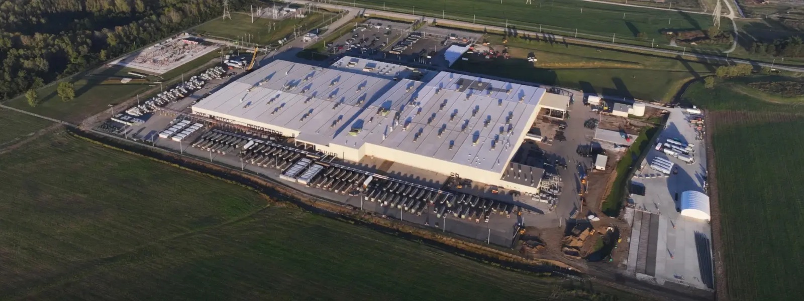 Aerial photograph of an xAI Colossus data center campus: a warehouse-scale building with white roofs, long rows of power and cooling machinery along its sides, a substation at one corner, and open fields around it.