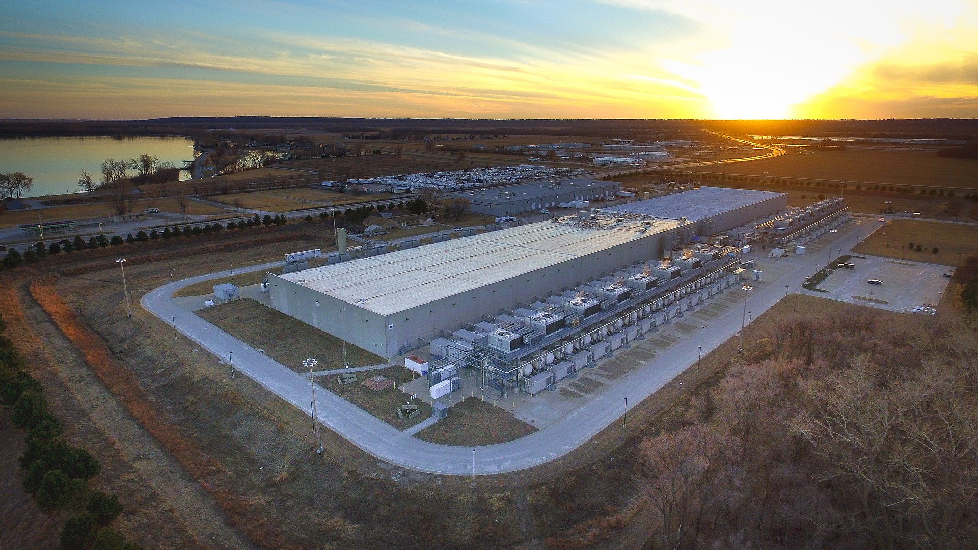 Aerial photograph at dusk of a Google data center outside Council Bluffs, Iowa: one long warehouse-scale building with rows of cooling equipment along its wall, surrounded by roads, fields, and a lake.