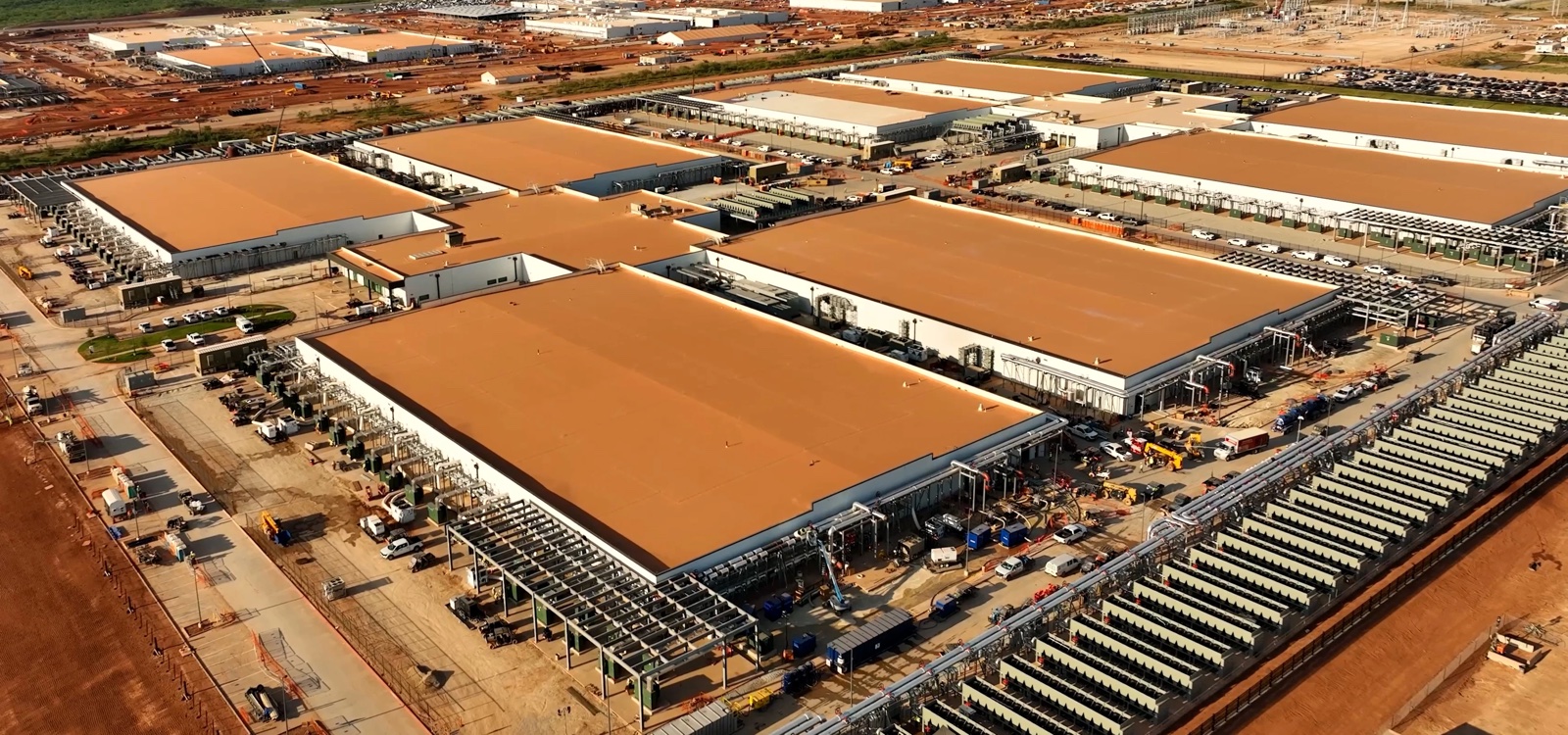 Aerial photograph of OpenAI's Stargate campus under construction outside Abilene, Texas: rows of warehouse-scale data halls with tan roofs, cooling infrastructure lining their walls, and construction equipment across the site.